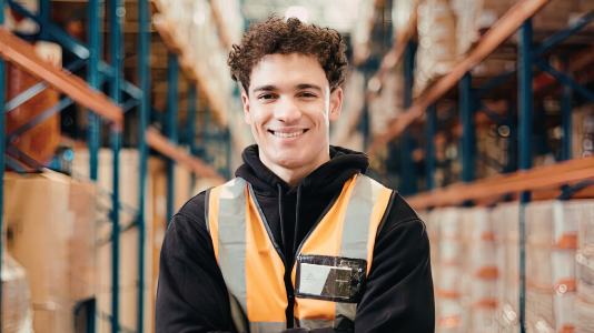 A young worker in a safety vest smiling in a warehouse, representing ProServeIT's IT services for the manufacturing industry