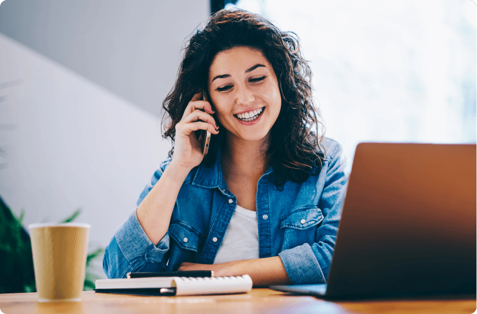 A woman smiling while on a phone call at her desk with a laptop open in front of her, representing the seamless IT operations and responsive support ProServeIT provides