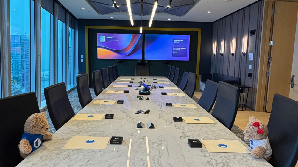 Hope and Hugo teddy bear mascots at a conference room setup for a ProServeIT AI Executive Briefing Session (EBS) in Toronto, with presentation screens, meeting materials, and city skyline views.